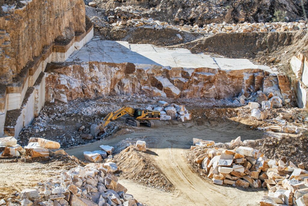 An excavator operates in a large stone quarry with rugged rocky terrain under daylight.